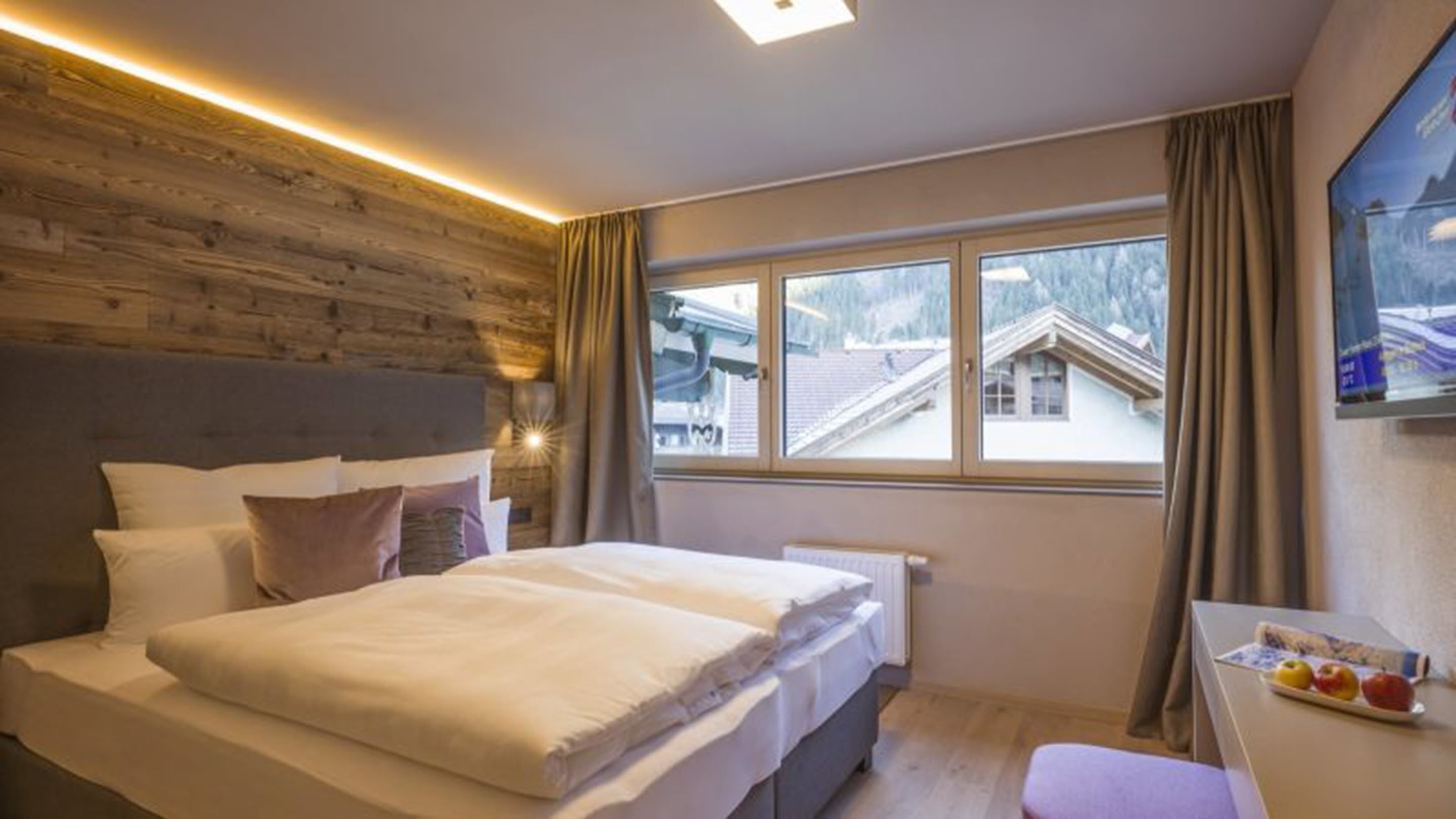 Une chambre à coucher avec une tête de lit en bois, un lit recouvert de draps blancs, une fenêtre donnant sur des maisons et une petite table sur laquelle se trouve des fruits.