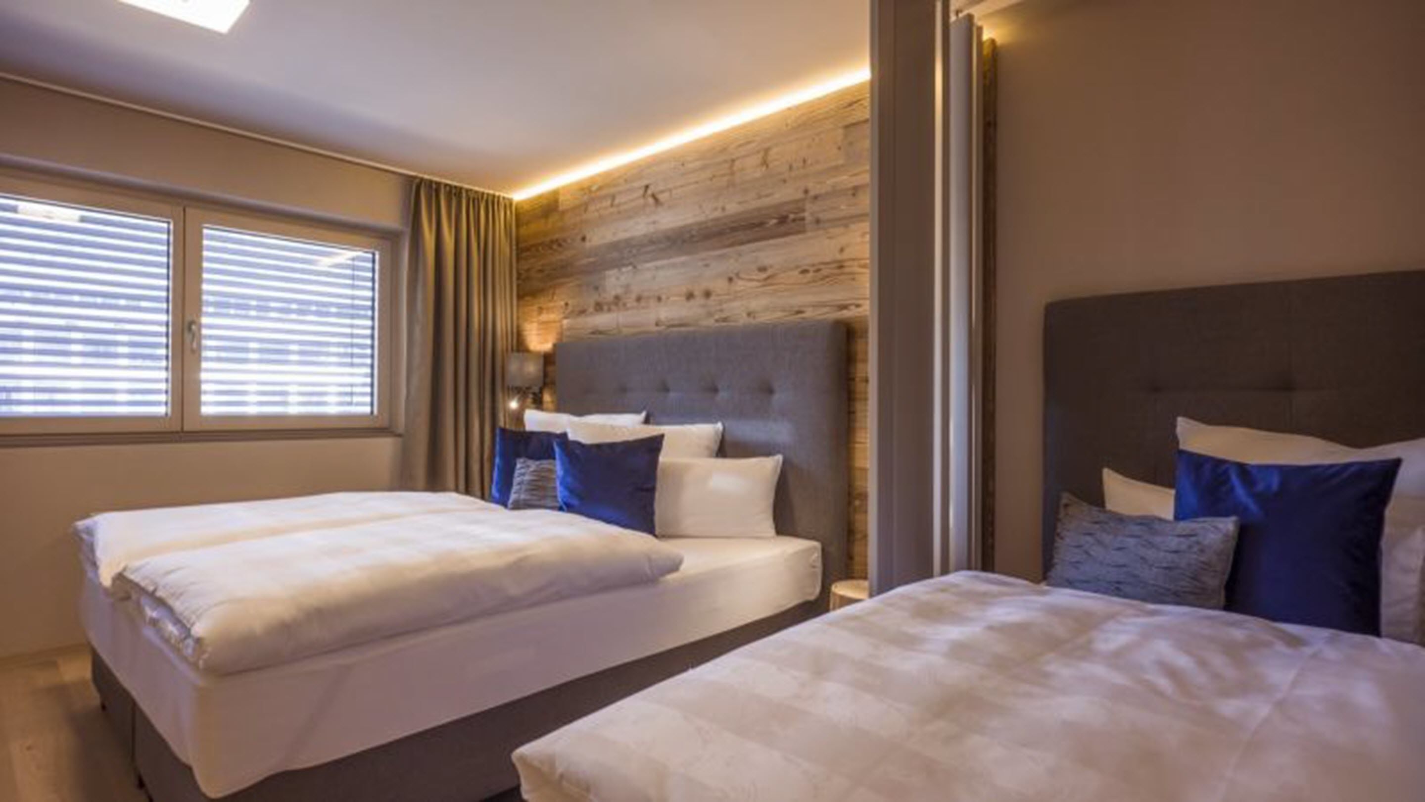 A hotel room with two beds, a wooden accent wall, and a window with blinds.