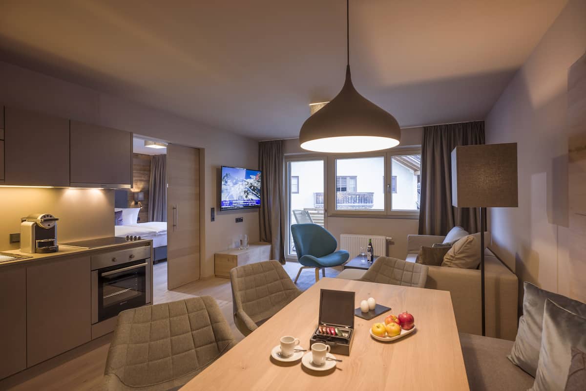 A modern hotel room with a kitchenette, a dining area with a table set for two, a blue armchair, and a large window with a city view.
