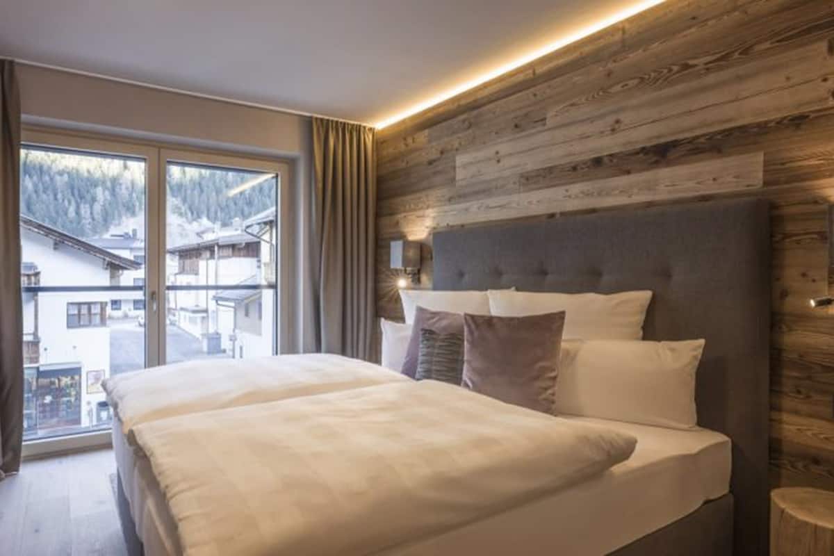 A bedroom with a large bed, wooden panel wall, and a view of buildings through a window.
