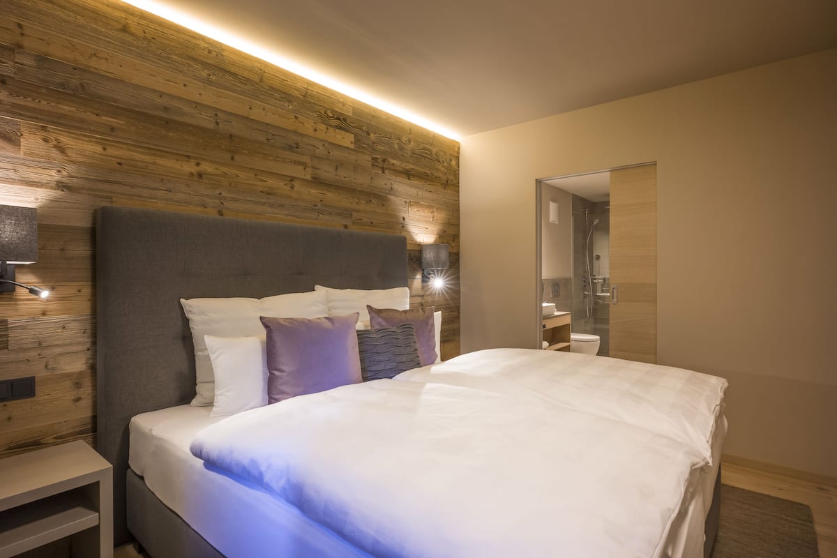 A modern bedroom with a wooden headboard, a large bed with white and purple bedding, and a bedside table with a lamp.