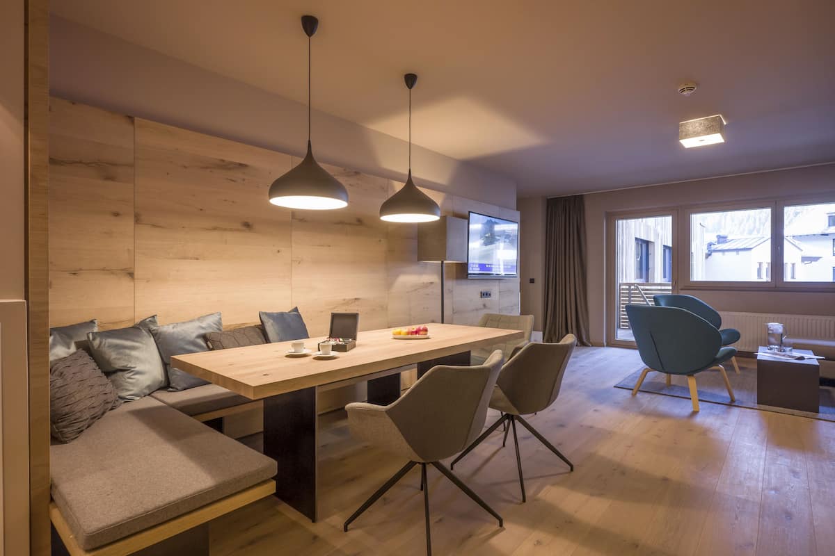 A modern dining area with a wooden table, grey upholstered seating, pendant lighting, and a view of the outside through large windows.