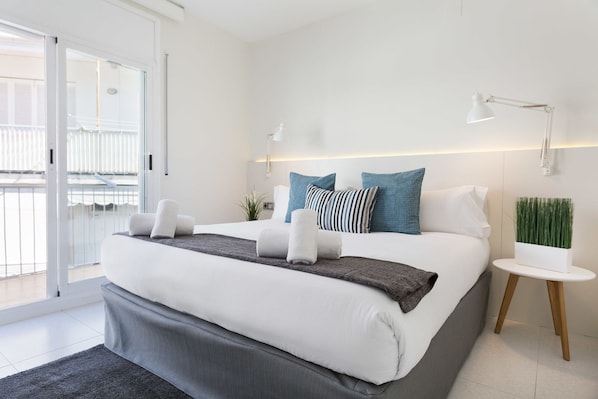 A modern bedroom with a large bed, a small bedside table, a potted plant, and a view of the balcony.