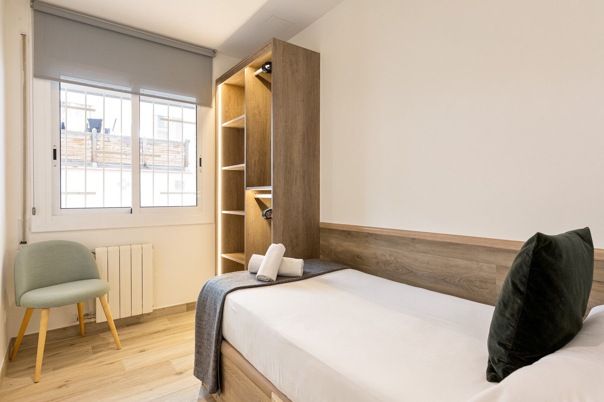 A bedroom with a bed, a chair, a wardrobe, and a window with a view of the outside.