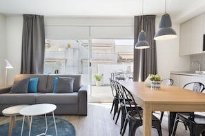 A modern living room with a grey sofa, two white side tables, a wooden dining table with black chairs, and a kitchen area in the background.