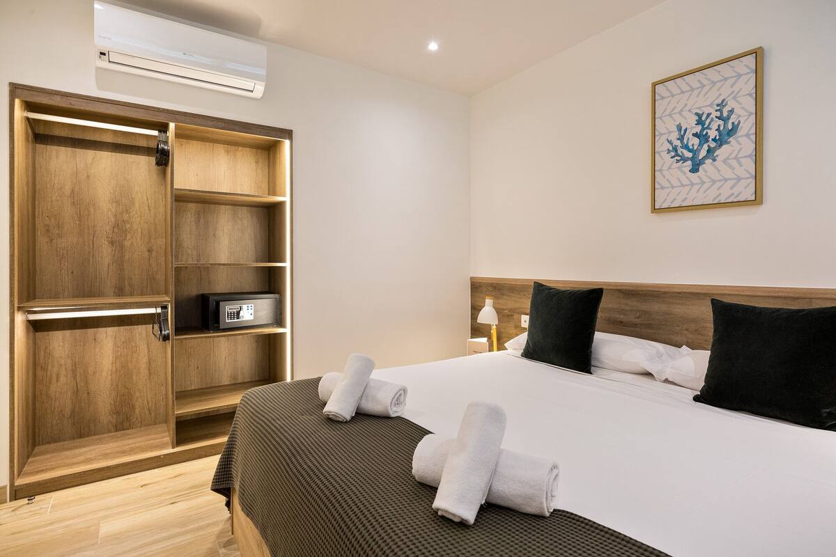 A hotel room with a bed, a wooden wardrobe, a wall-mounted air conditioner, and a framed artwork.