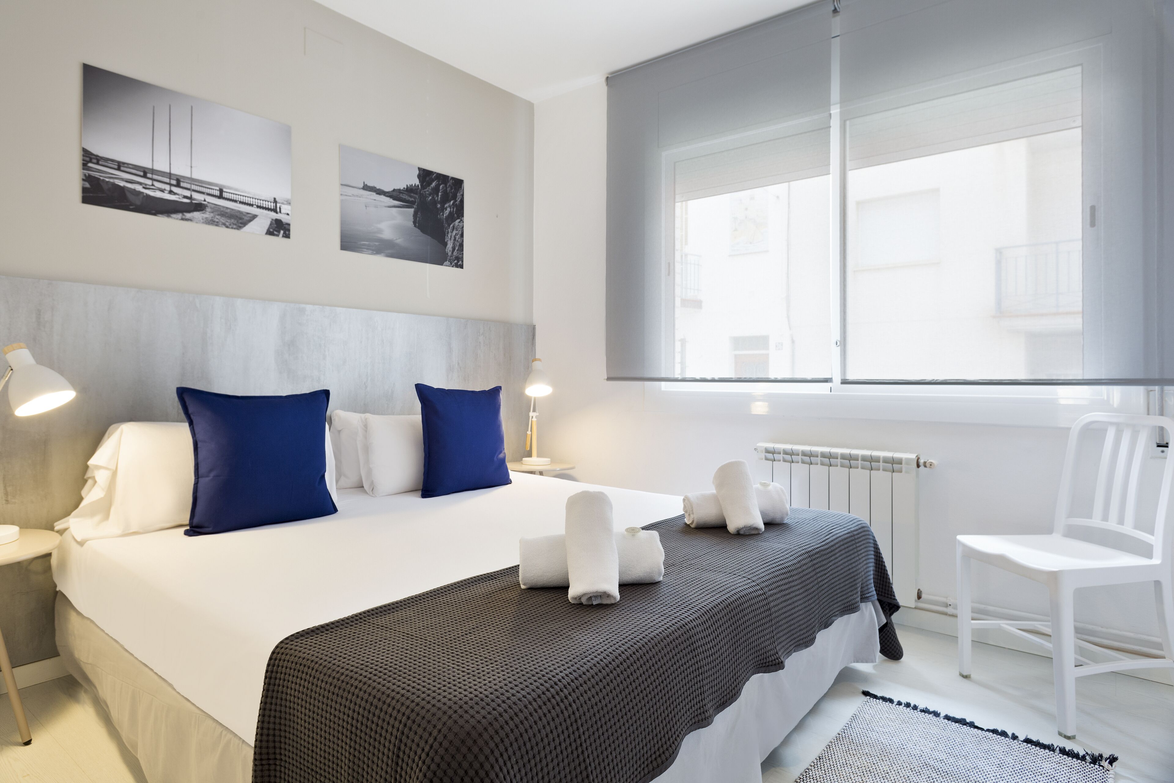 A modern hotel room with a large bed, two blue pillows, a grey headboard, a white chair, and a window with a view of buildings.