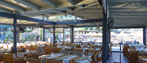 Un restaurante al aire libre con mesas y sillas de madera, techo cubierto y vista a la playa.