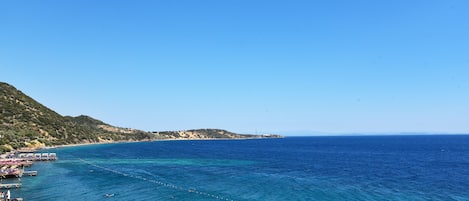 Vista costeira com mar azul cristalino, guarda-sóis de praia e montanhas ao fundo.