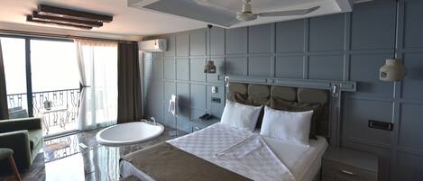 A modern hotel room with a large bed, a round bathtub, a balcony with a view, and a ceiling fan.
