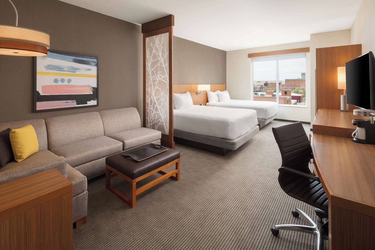 Habitación de hotel con sofá, dos camas, escritorio, televisor y ventana con vista a la ciudad.