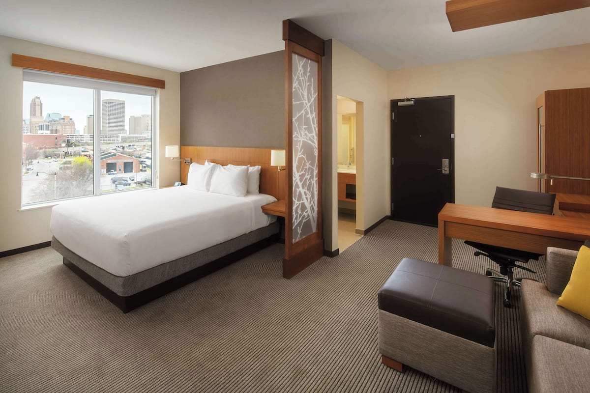 Habitación de hotel con una cama grande, un escritorio, una silla y vistas a la ciudad a través de un ventanal amplio.