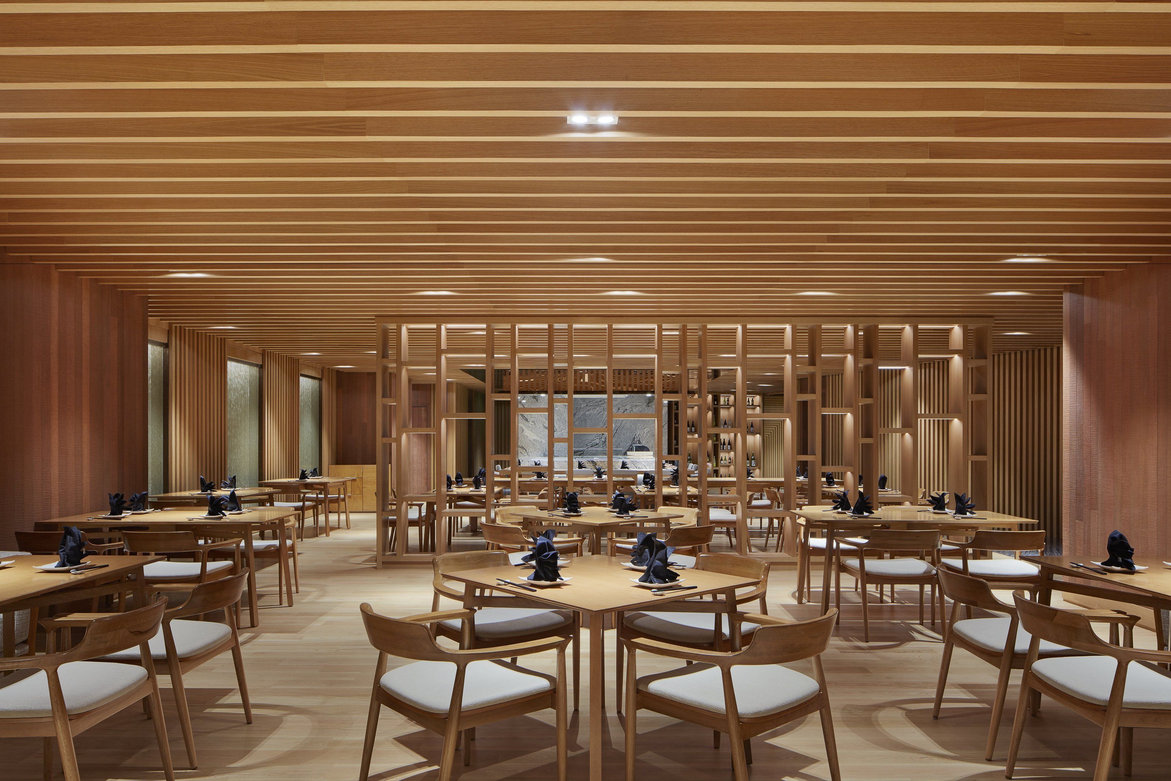 Un vaste espace dédié à la salle à manger, avec de nombreuses tables et chaises en bois, une structure décorative centrale également en bois, et de grandes fenêtres qui laissent entrer la lumière naturelle.