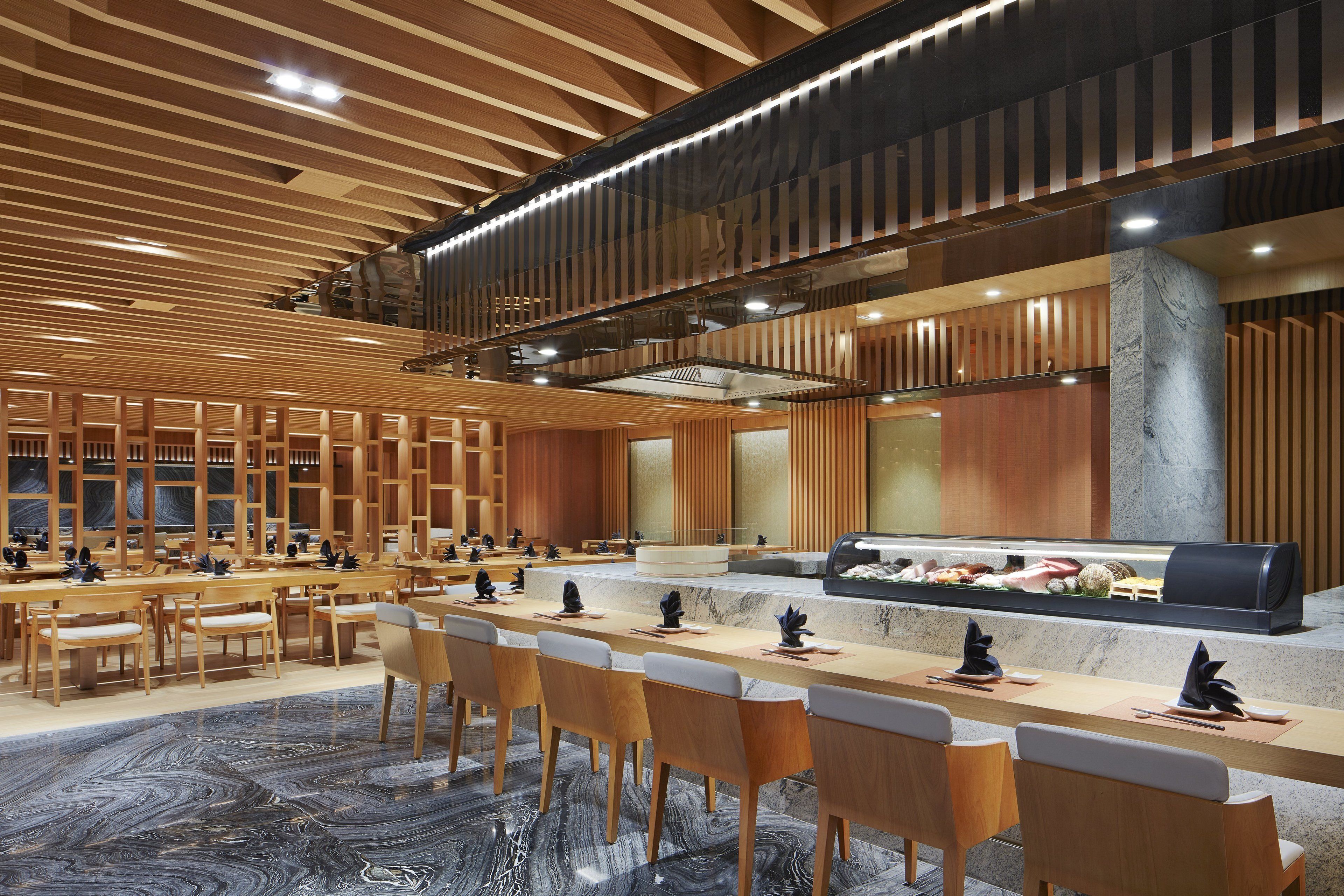 Un hall d’hôtel spacieux, doté d’un long comptoir d’accueil, de plusieurs espaces salon et d’un design moderne caractérisé par des panneaux en bois et un sol en marbre.