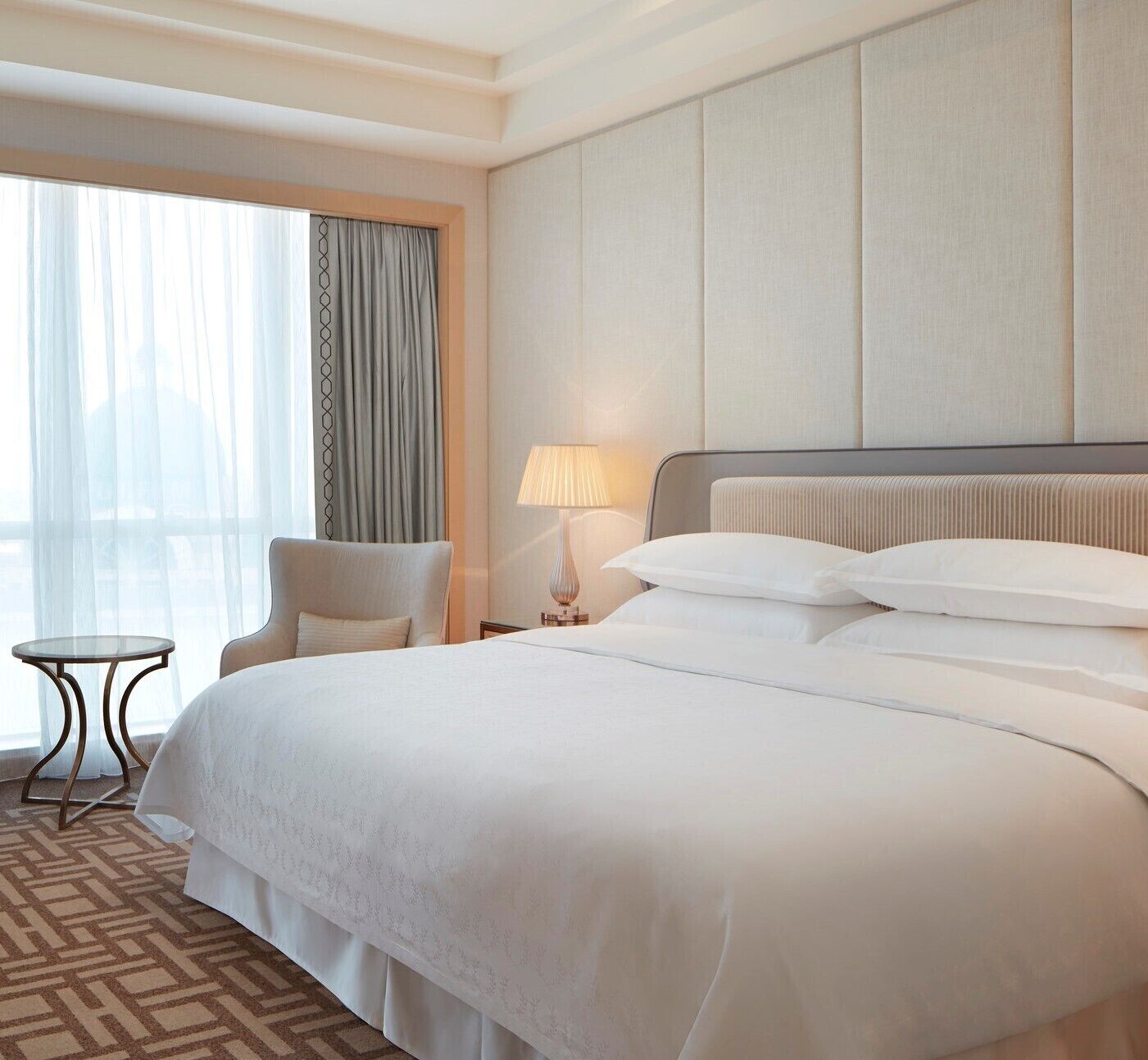 A hotel room with a large bed, a chair, a small table, and a lamp.