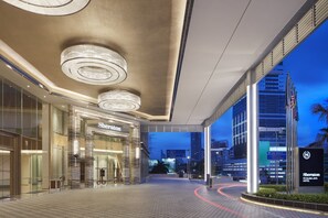 Entrada de hotel con diseño moderno, fachada de vidrio, un cartel llamativo del hotel y vista del paisaje urbano al fondo.