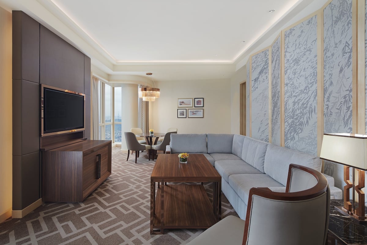 A modern hotel room with a large flat-screen TV, a wooden coffee table, a grey sofa, and a dining area with a chandelier.