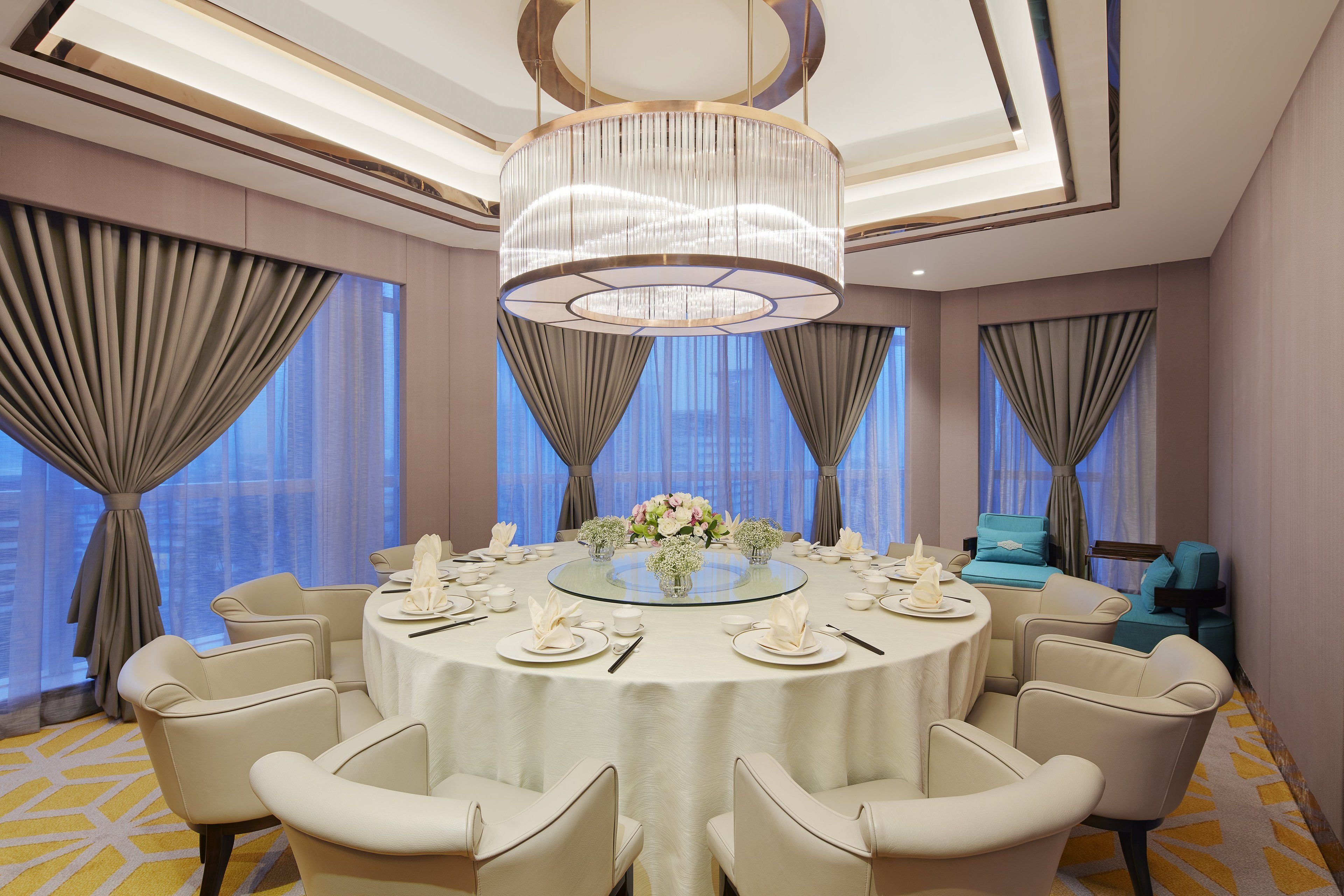 A round dining table with white tablecloth and chairs, surrounded by curtains and a chandelier.