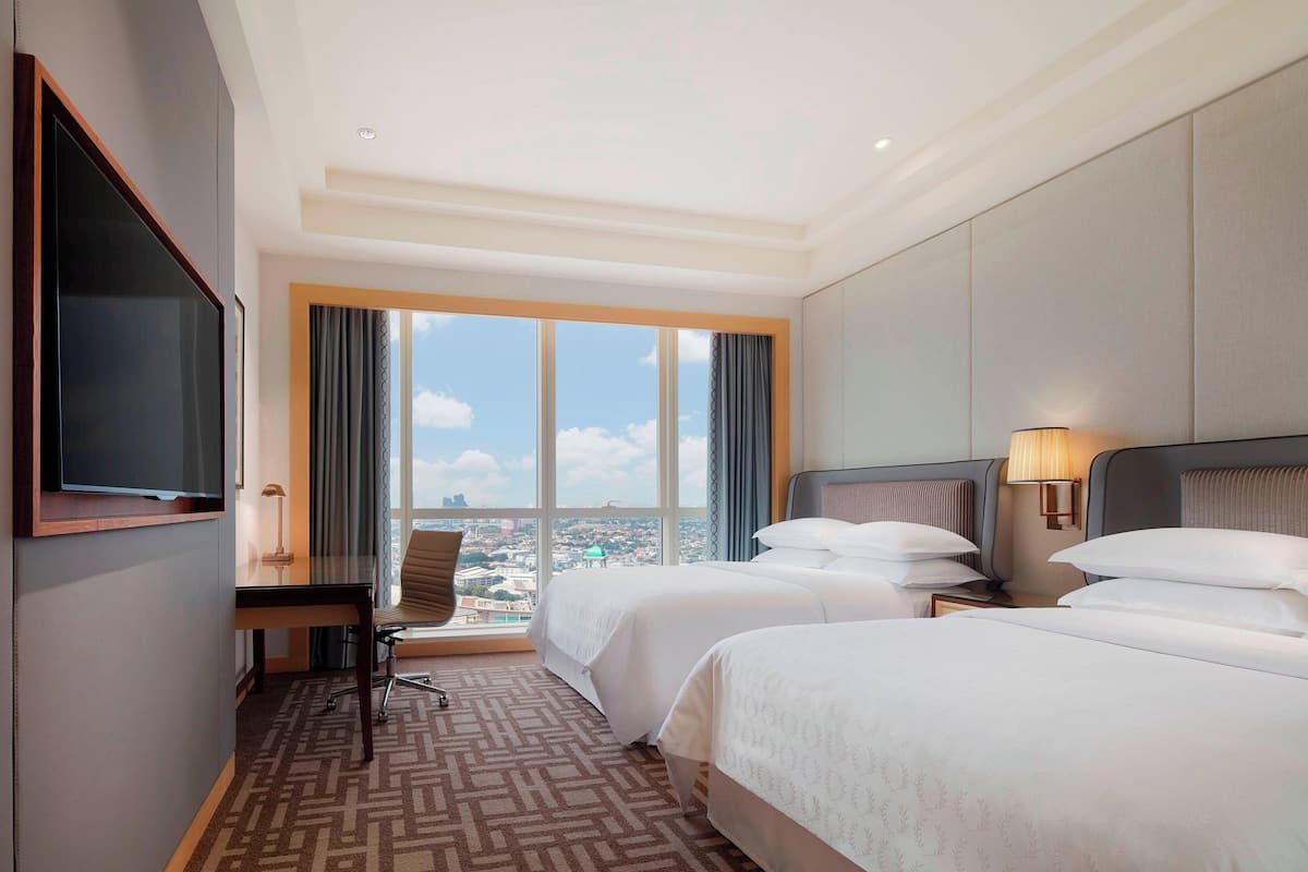A hotel room with two beds, a desk, a chair, a TV, and a large window offering a city view.