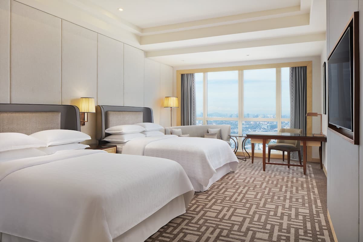 A hotel room with two beds, a desk, a chair, and a large window offering a city view.