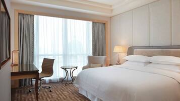 A hotel room with a large bed, a desk with a chair, a small table, and a large window with curtains.