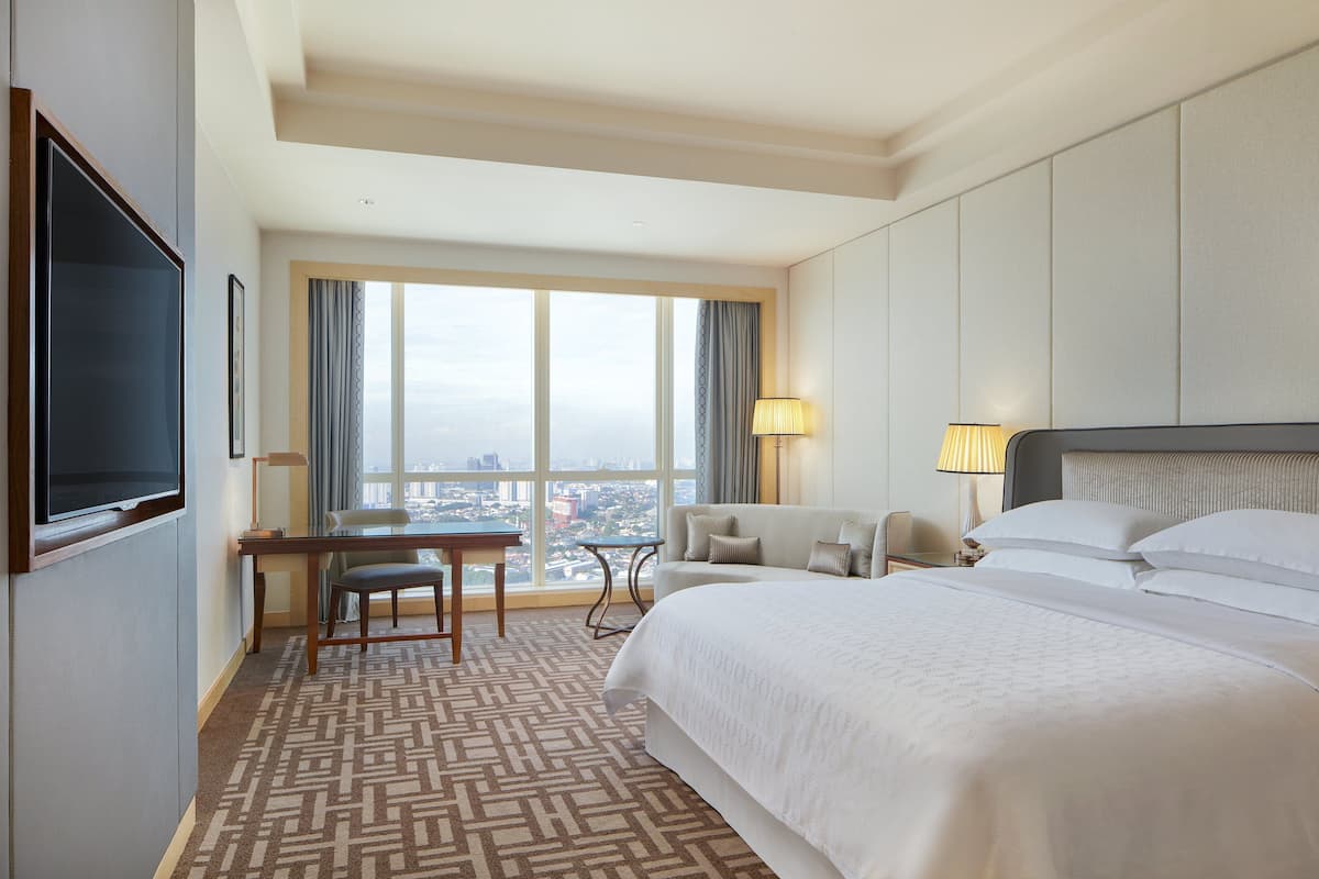 A hotel room with a large bed, a desk, a chair, a TV, and a view of the city through large windows.