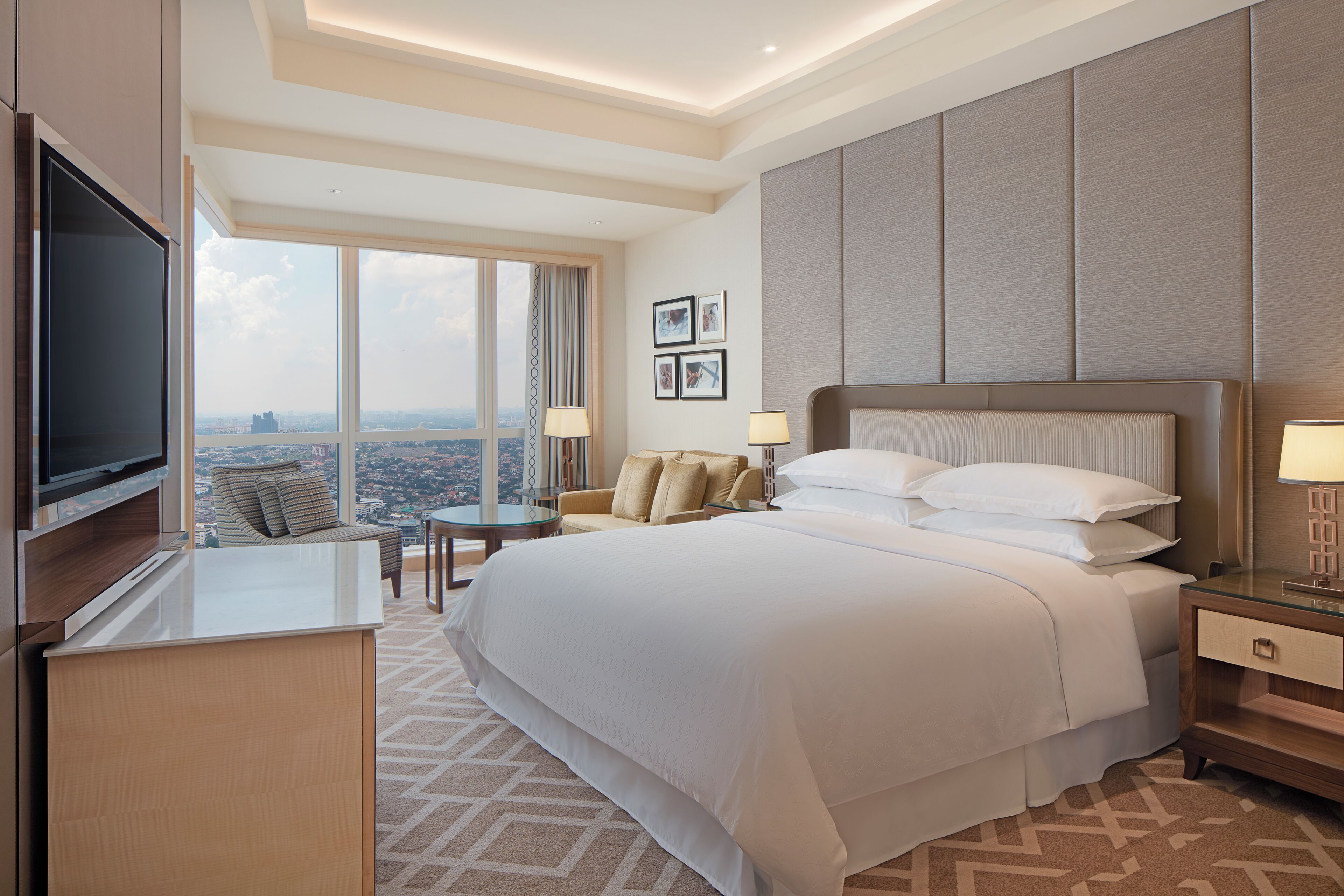 A hotel room with a large bed, a TV, a seating area with a view, and a cityscape visible through the window.