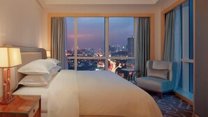 A hotel room with a large bed, a chair, a nightstand, and a view of a cityscape at dusk.