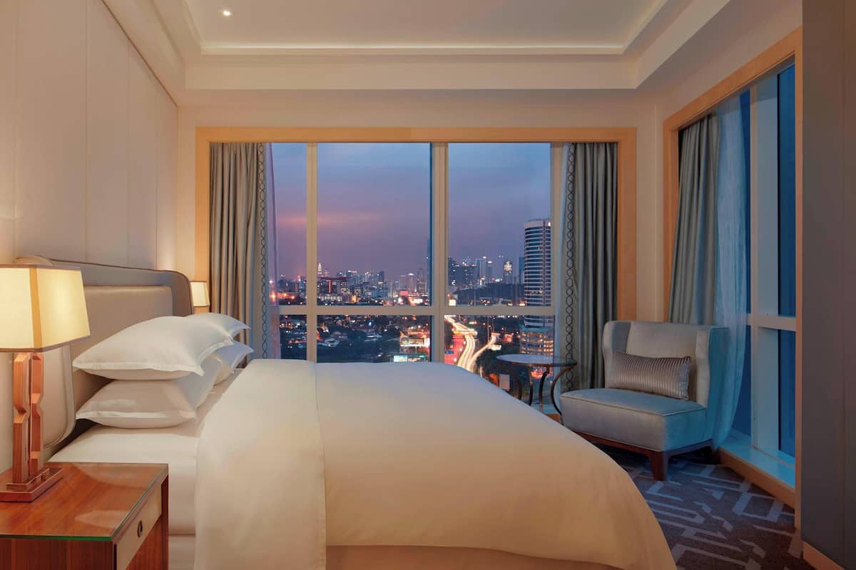 A hotel room with a large bed, a chair, a nightstand, and a view of a cityscape at dusk.