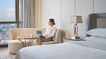 A hotel room with a bed, a sofa, a small table with a laptop and a cup, a lamp, and a view of a cityscape through large windows.