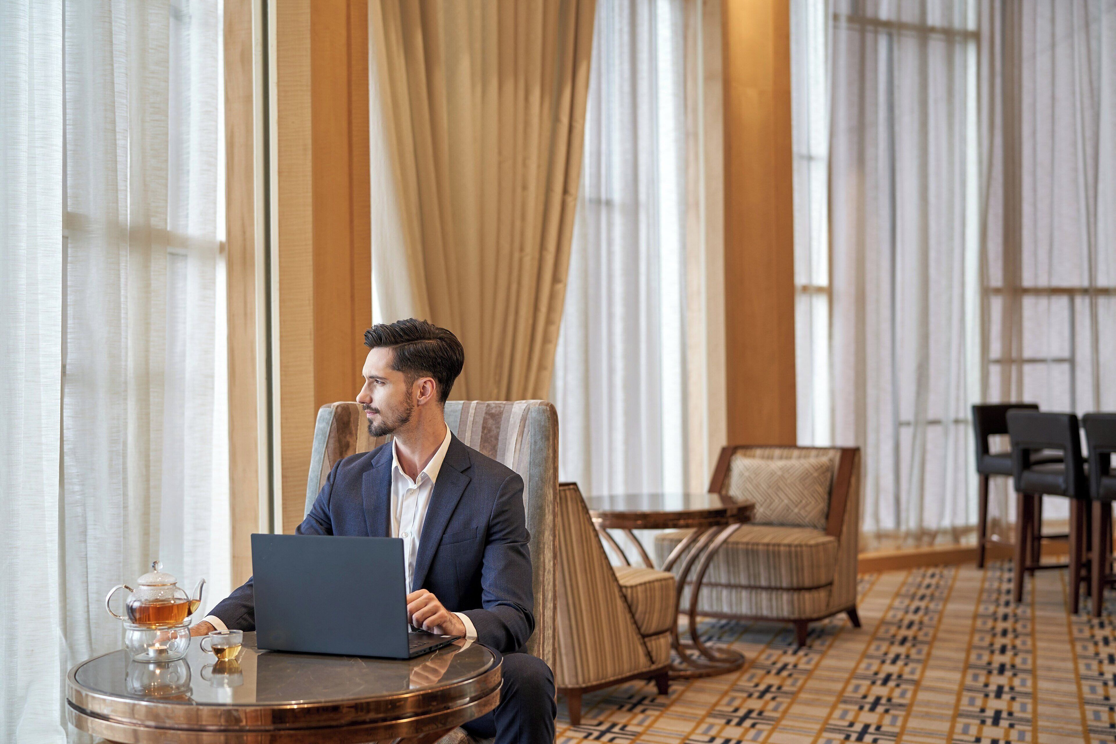Ein Mann in einem Anzug bedient einen Laptop in einem Hotelzimmer. Es sind ein runder Tisch, ein Stuhl und ein gemustertes Teppich zu sehen.