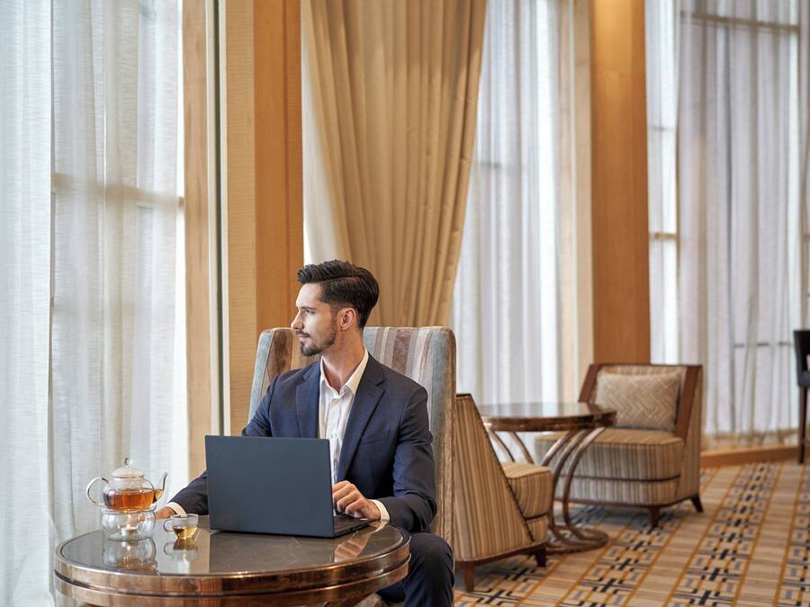 Ein Mann in einem Anzug bedient einen Laptop in einem Hotelzimmer. Es sind ein runder Tisch, ein Stuhl und ein gemustertes Teppich zu sehen.