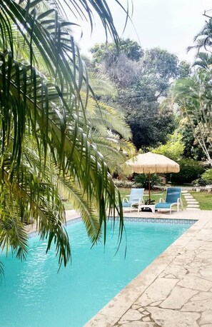 Piscina con sillas de descanso y una sombrilla de paja, rodeada de exuberante vegetación.