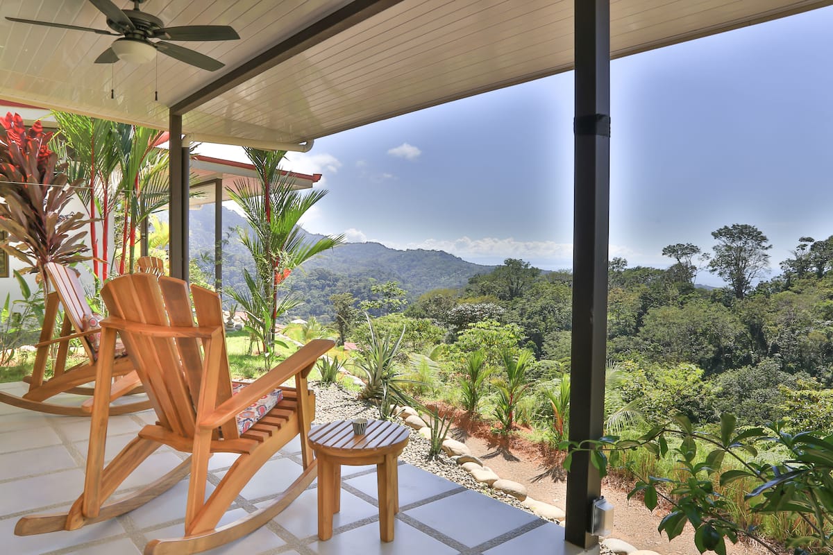 A covered patio with wooden rocking chairs and a small table, overlooking a lush garden and distant mountains.