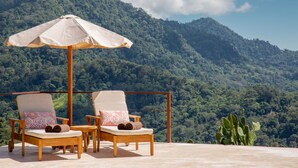 Dos sillones de madera con cojines y un paraguas color beige, frente a imponentes montañas cubiertas de vegetación.