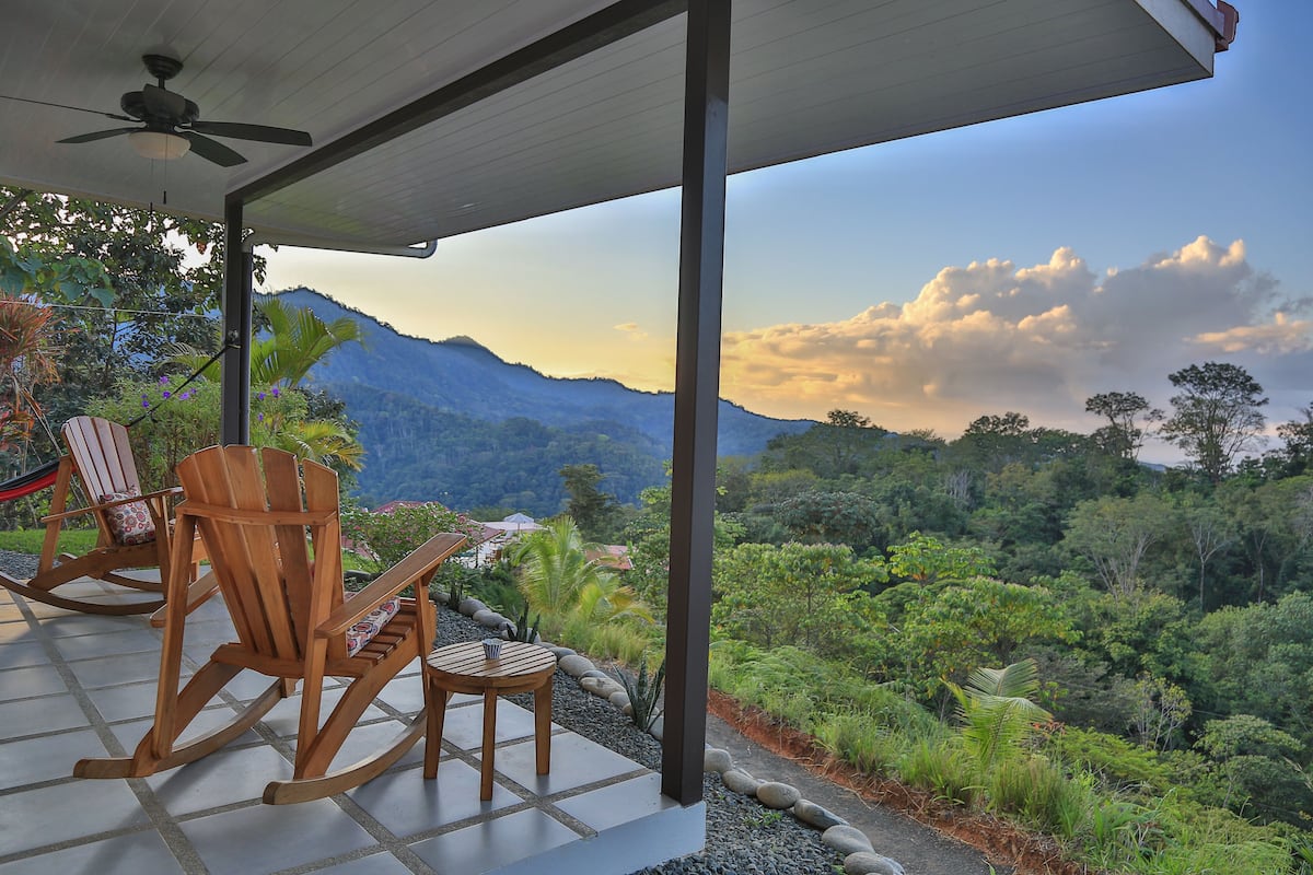 A covered patio with wooden rocking chairs and a table, overlooking a lush mountainous landscape.