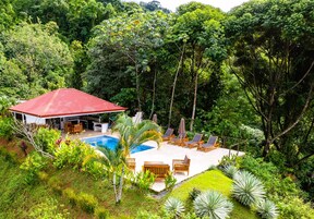 Una casa con techo rojo, piscina y patio con sillones de descanso.