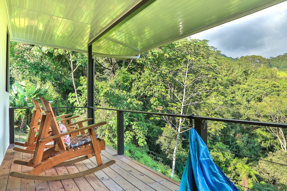 A wooden rocking chair on a wooden deck with a view of a lush forest.