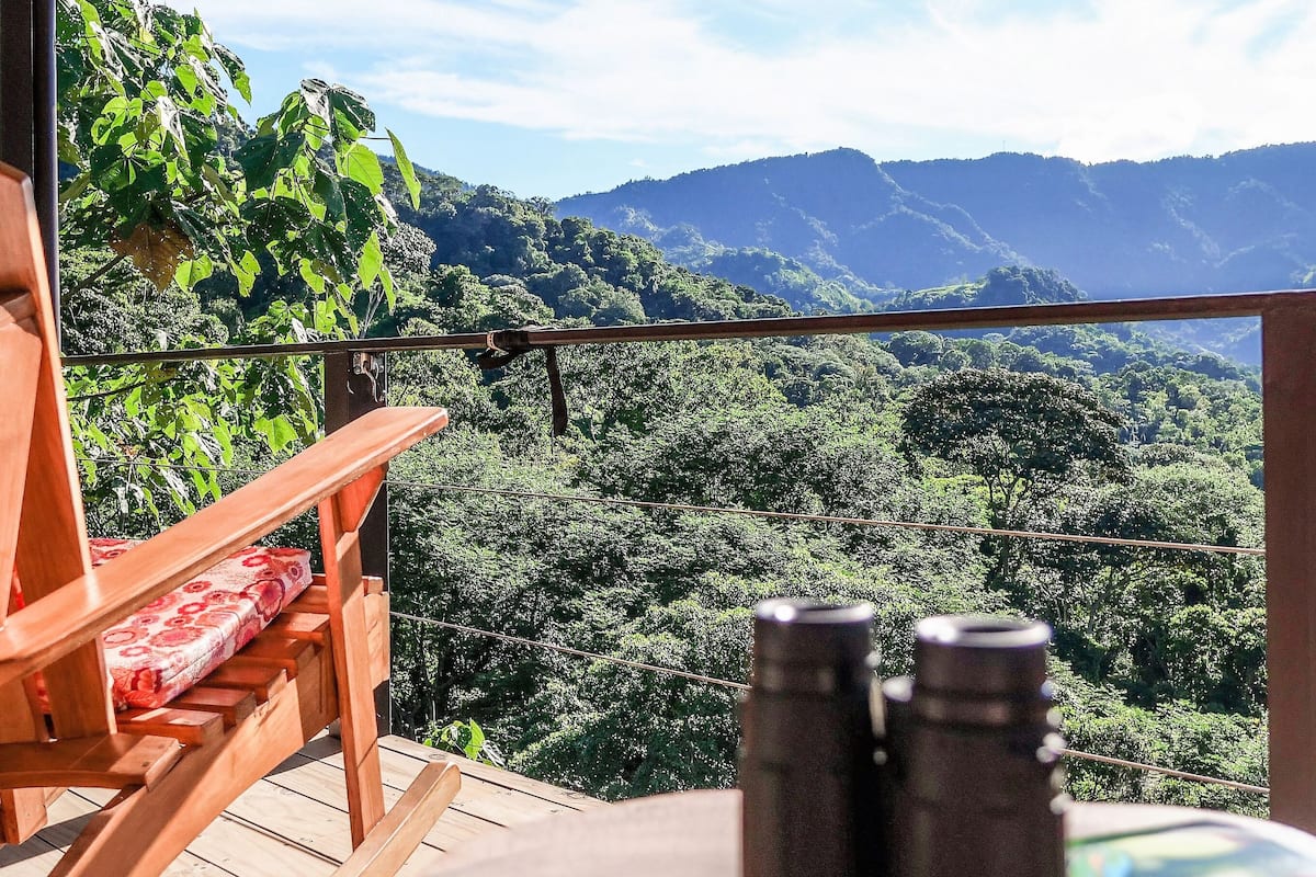 A wooden deck with a chair and binoculars, overlooking a lush forest and mountains.