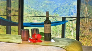 Una mesa con una botella de vino, dos copas y una flor roja en un balcón con vistas a un paisaje montañoso.