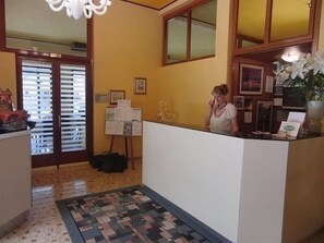 A reception area with a receptionist, a bulletin board, a plant, and a patterned rug. - Mayer (Jesolo)