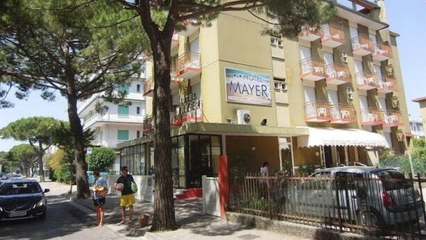 Un hôtel appelé « Hôtel Mayer », avec une enseigne, de nombreux balcons et un espace de stationnement à l’avant.