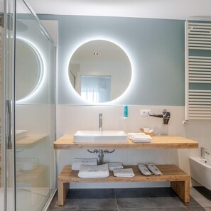 A modern bathroom with a glass shower enclosure, a wooden vanity with a sink, a round mirror, and a built-in bench with towels. - Mayer (Jesolo)