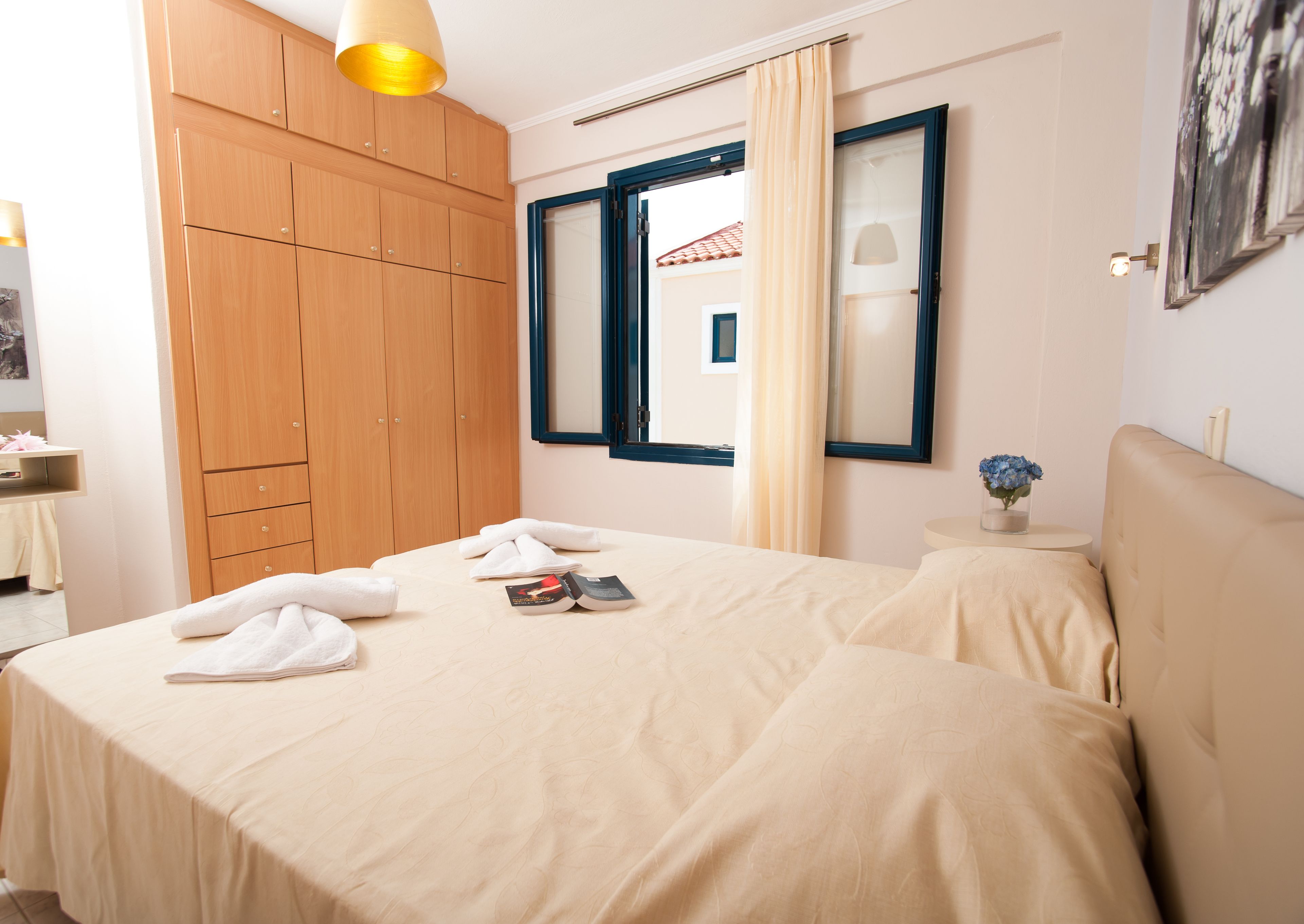 A hotel room with a bed, a wooden wardrobe, a mirror, a window with curtains, a bedside table with a vase of flowers, and a wall-mounted lamp.