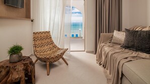 Ein Hotelzimmer mit großem Fenster, einer Couch, einem Sessel, einem an der Wand montierten Fernseher und einer kleinen Pflanze auf einem Holzschemel.