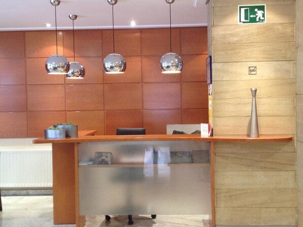 A reception desk with a wooden panel wall, three pendant lights, a green emergency exit sign, and a silver vase on a shelf. - Hotel Teremar (Benidorm)