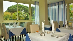 A dining area with tables set for two, large windows with curtains, and a view of greenery outside. - Helios (Cervia)