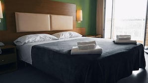 A hotel room with a large bed, a wooden headboard, a nightstand, a lamp, and a window with a view. - Helios (Cervia)