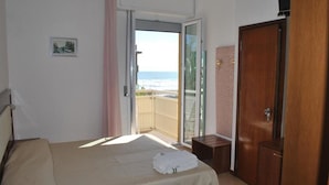 A bedroom with a bed, a wooden wardrobe, a small bench, and a sliding glass door leading to a balcony with a view of the sea. - Helios (Cervia)