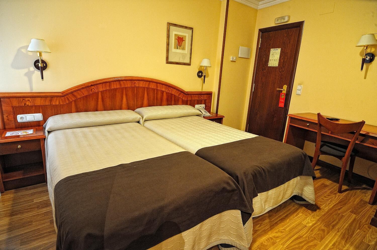 A double bed with a wooden headboard, two bedside tables with lamps, a desk with a chair, and a door with a sign.